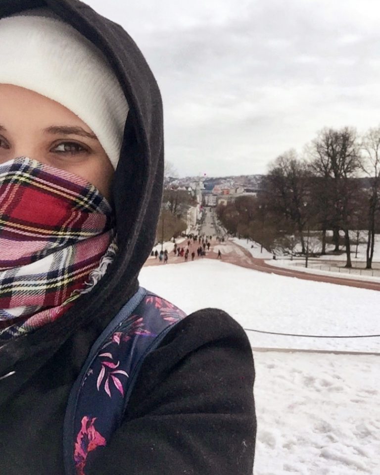 Rachel com gorro, cachecol cobrindo o rosto, casaco grosso com capuz, em rua com neve.