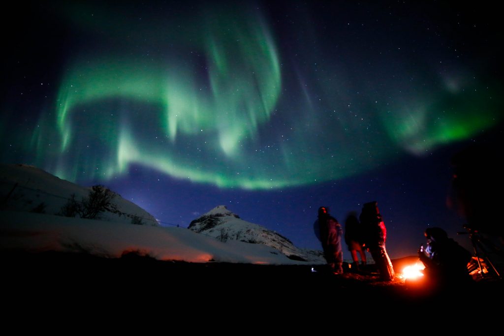 Foto da aurora boreal acima de uma montanha coberta de neve, durante viagem da Rachel para a Noruega. No canto inferior direito, pessoas estão ao redor de uma fogueira.