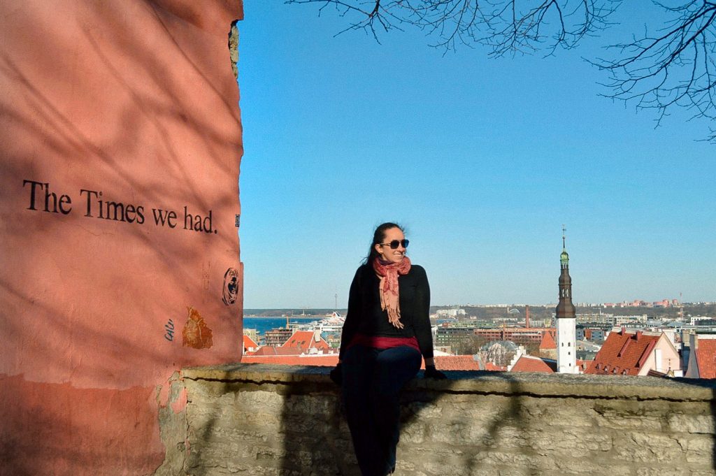 Rachel sentada em mureta ao lado de parede grafitada com a cidade de Tallin ao fundo.