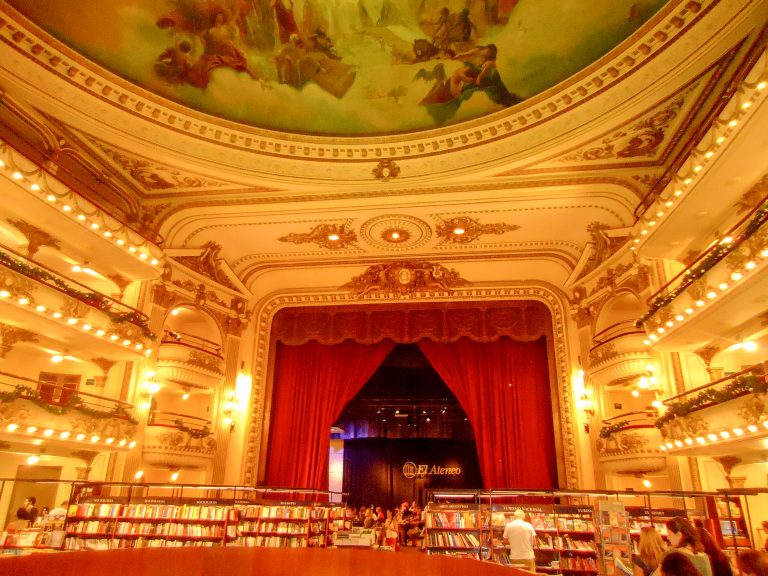 Teatro antigo com pinturas no teto, cortinas no palco ao fundo e prateleiras de livros.