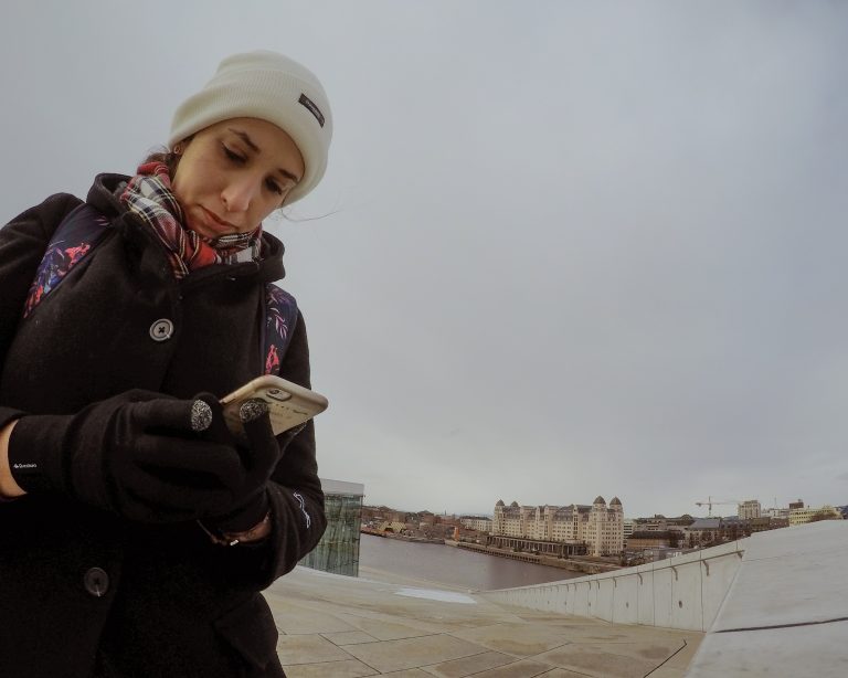 Rachel olha aplicativo no celular. Ao fundo, rio e prédios da cidade de Oslo.