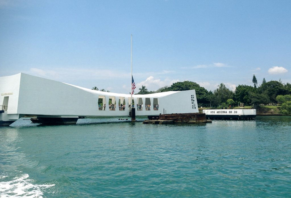 Memorial comprido sobre a água na baía de Pearl Habour, com visitantes nas janelas e bandeira dos EUA a meio-mastro.