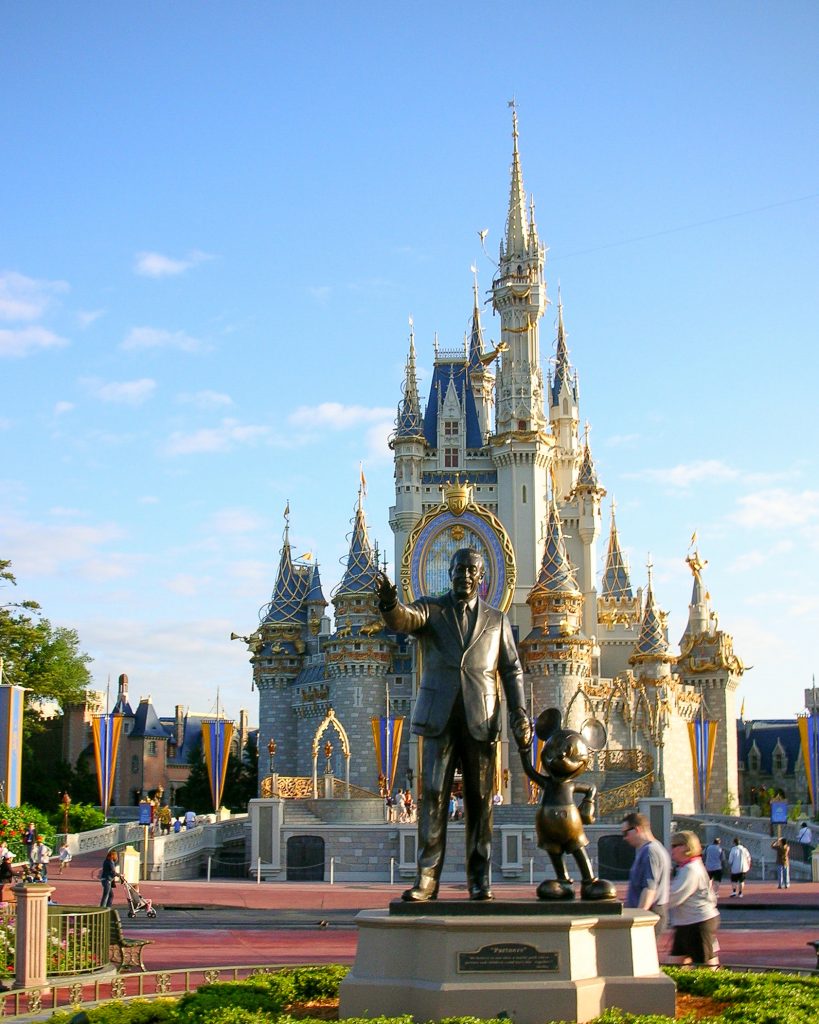 Estátua do Walt Disney de mãos dadas com o Mickey em frente ao castelo da Cinderela com detalhes dourados. Chão vermelho e céu azul.