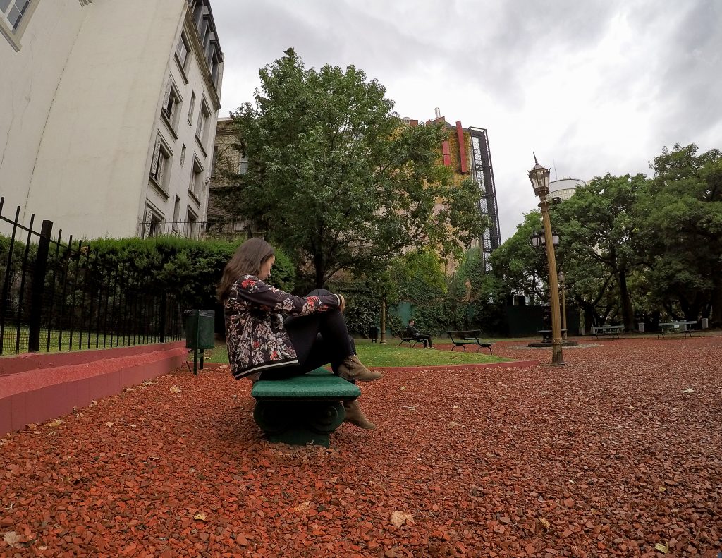 Rachel sentada em banco de praça com chão de cascalho e árvores ao fundo.